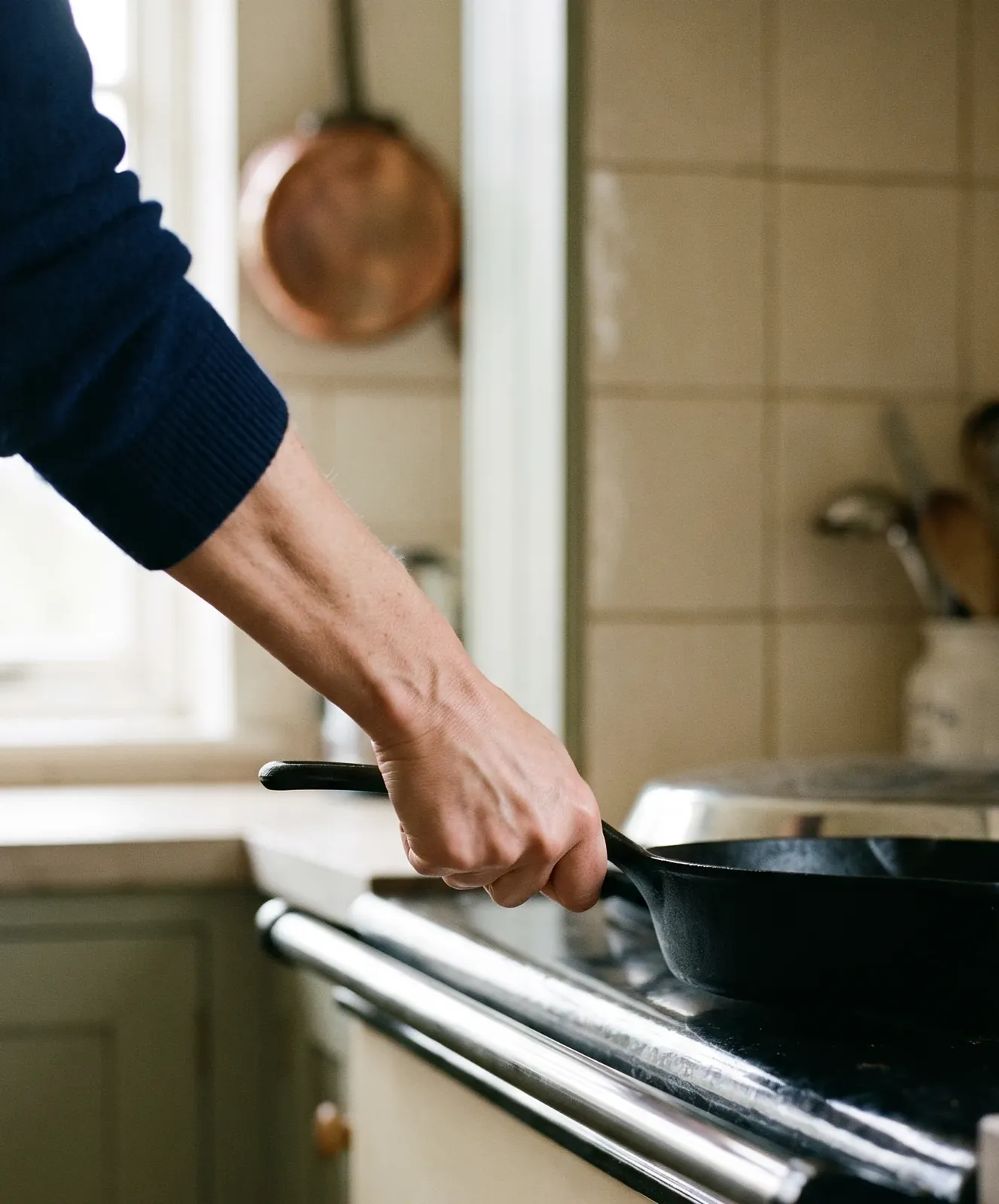 A hand lifting a heavy black cast-iron skillet off a cream range cooker, palm facing down