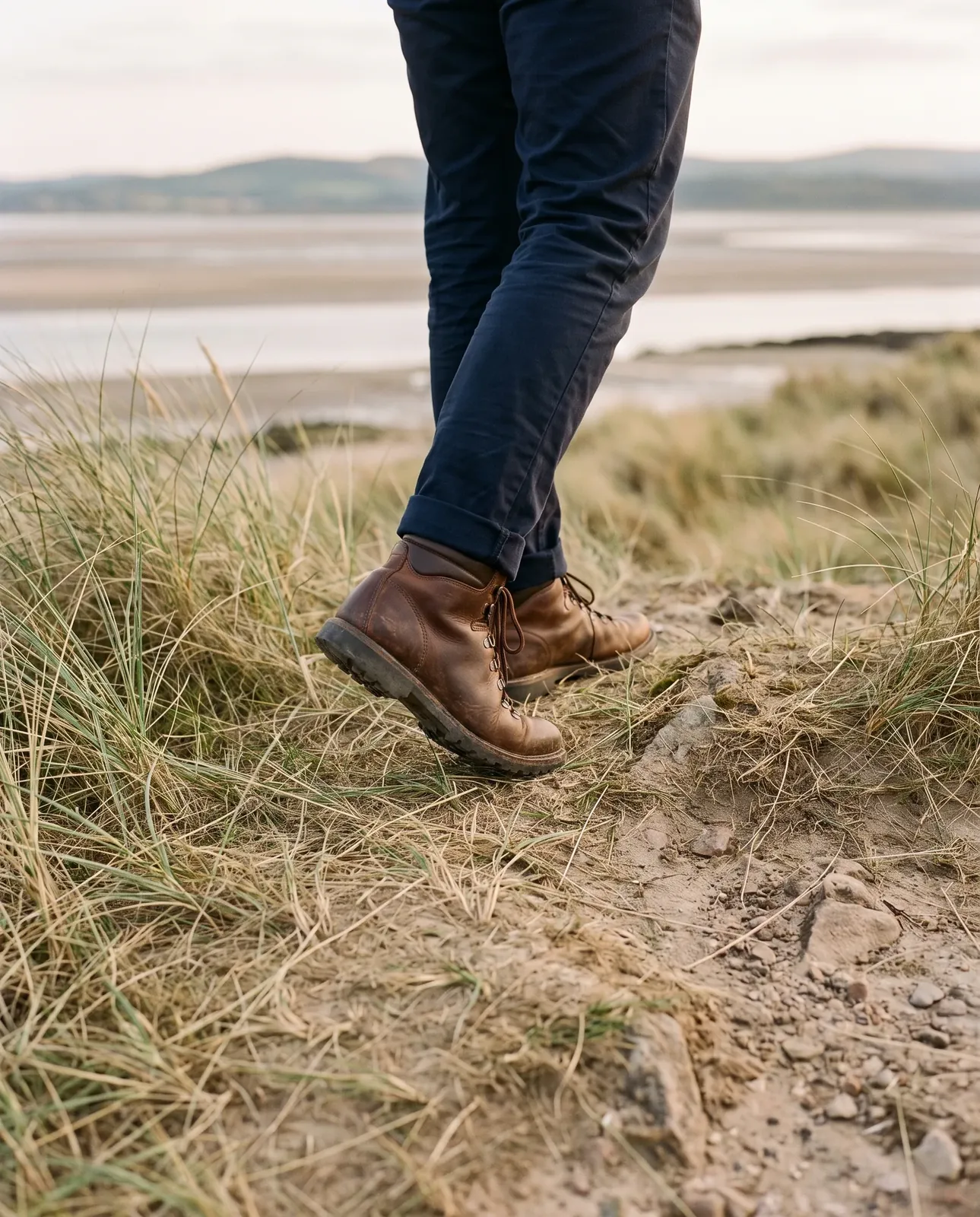 Brown leather walking boots stepping over an uneven, stony coastal path through sea grass