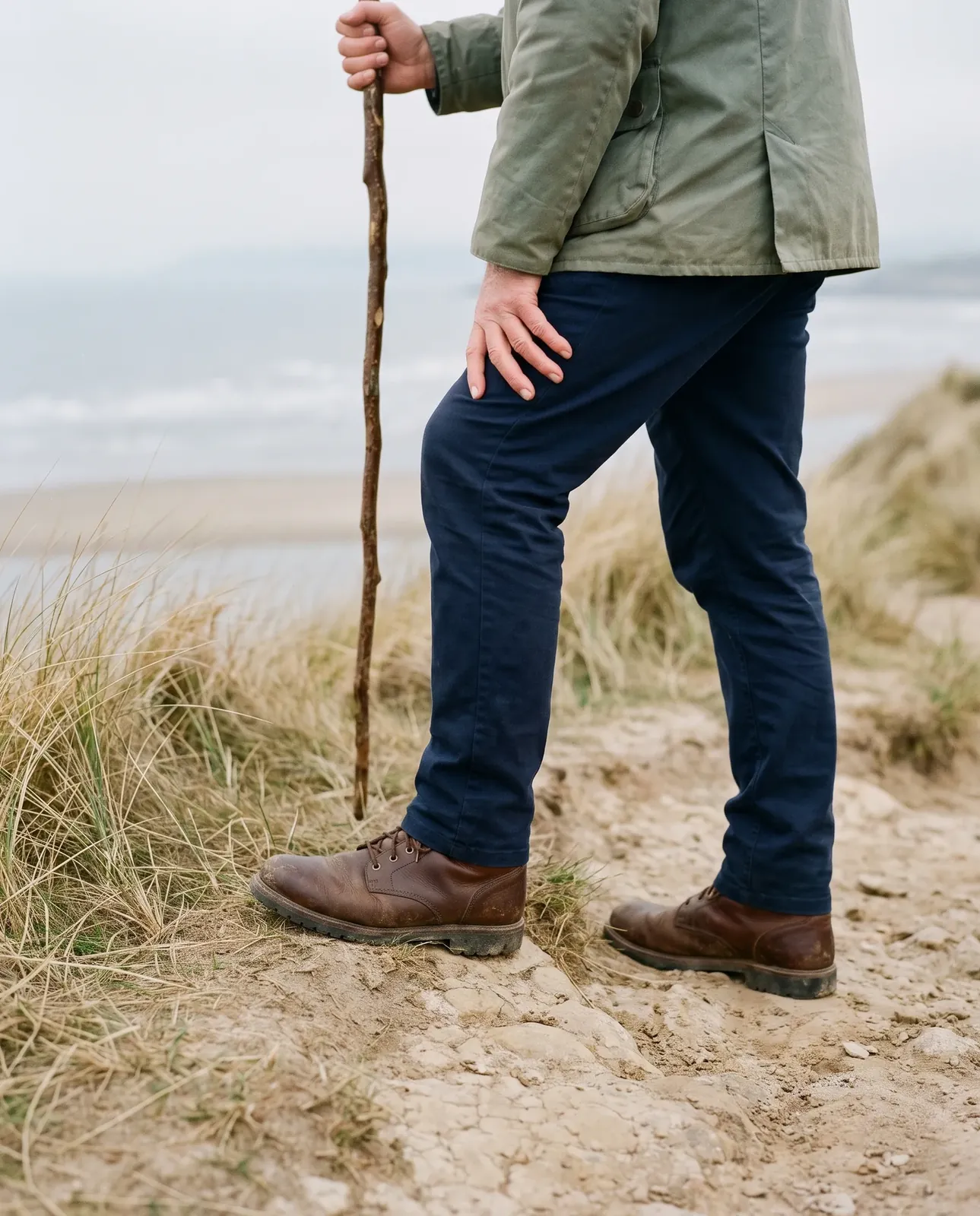 A walker pausing on a sandy coastal path, one hand on the knee