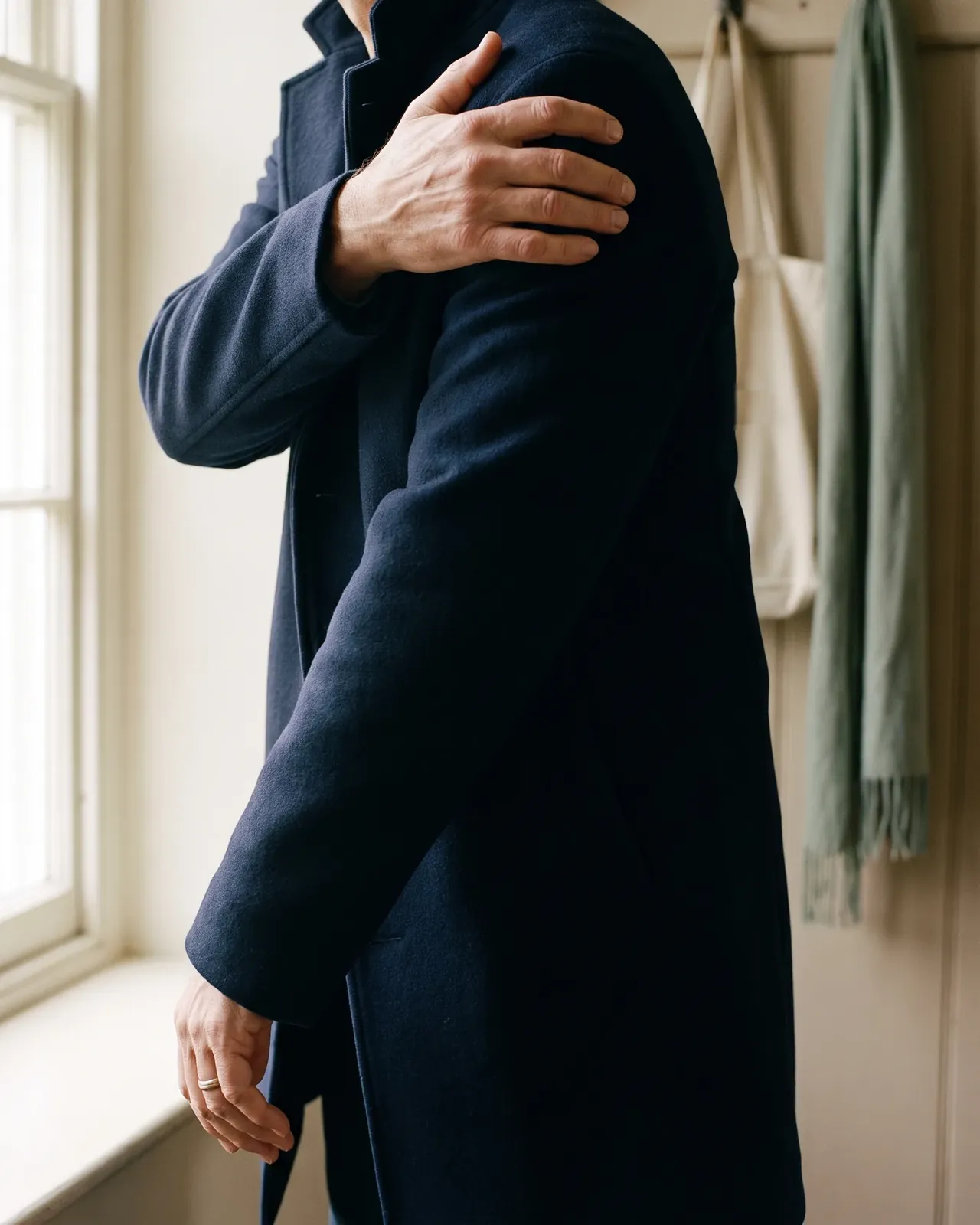 A man by a window in a navy coat, gripping the top of his painful shoulder as he dresses