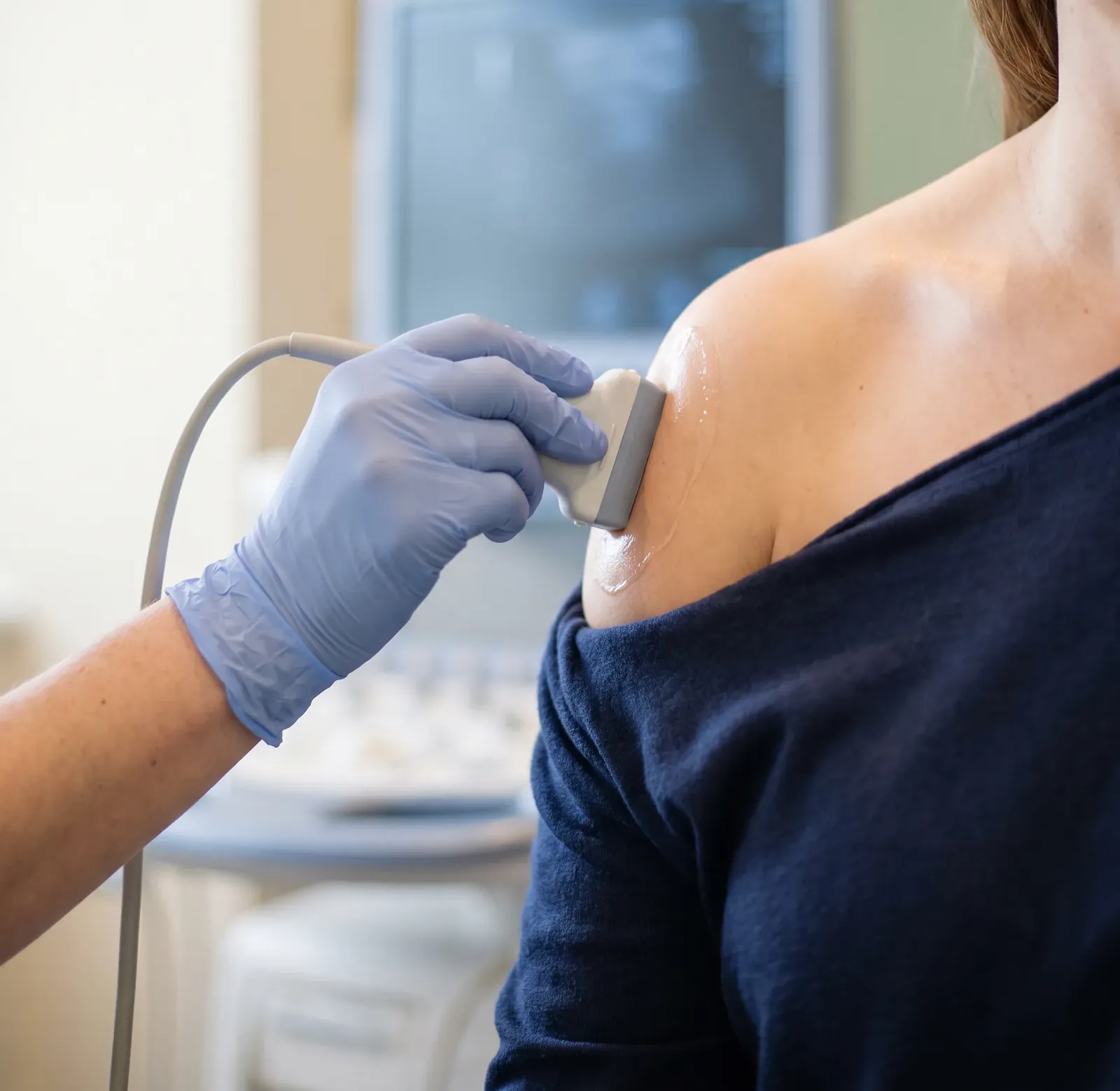 A gloved hand holding an ultrasound probe against a patient's shoulder, with the scanner screen behind
